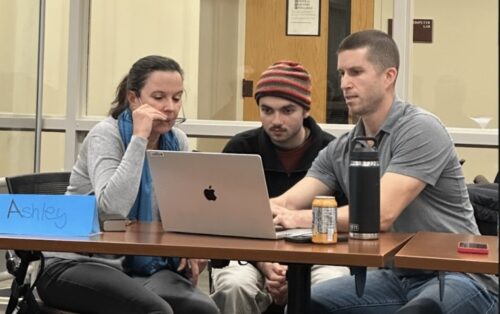 Three adults sit at a table focused on a laptop. A blue name card reading Ashley is in front, with drinks and a phone nearby. They appear to be in a classroom or office setting with glass walls and a door behind them.
