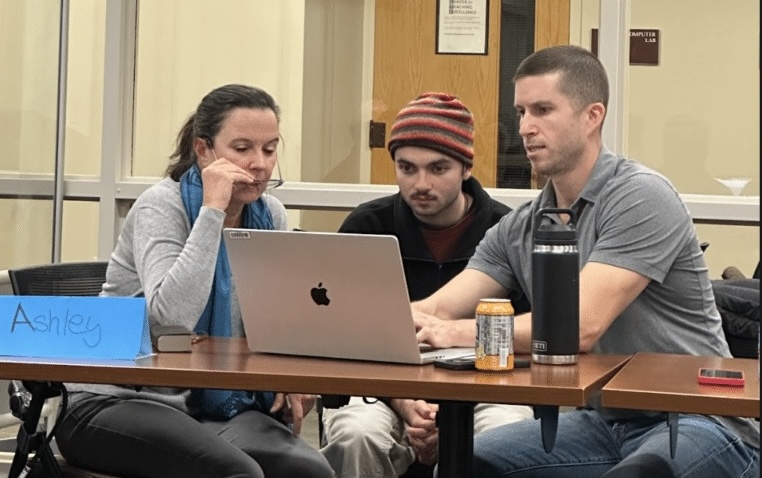 Three adults sit at a table focused on a laptop. A blue name card reading Ashley is in front, with drinks and a phone nearby. They appear to be in a classroom or office setting with glass walls and a door behind them.